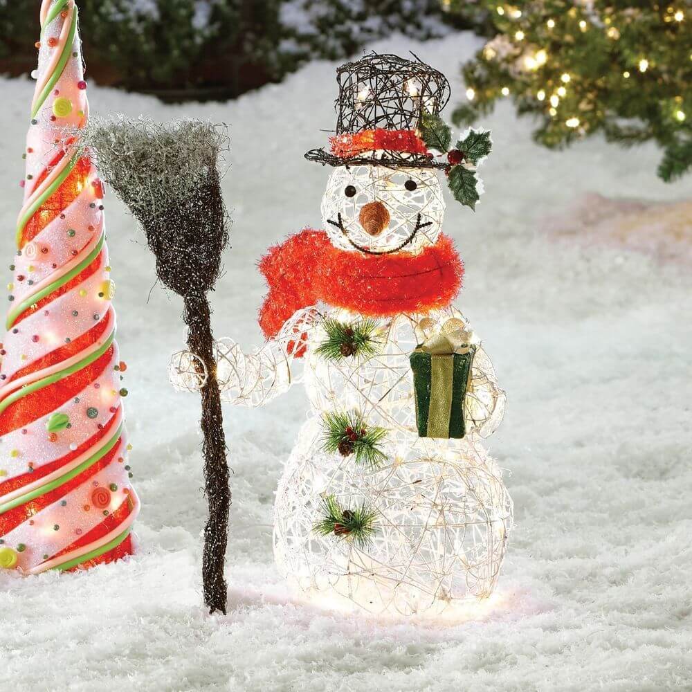 A snowman holding a broom next to a christmas tree
