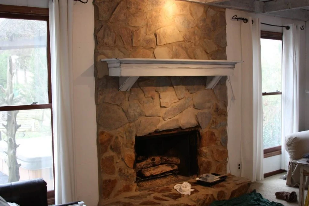 A stone fireplace in a living room next to a window
