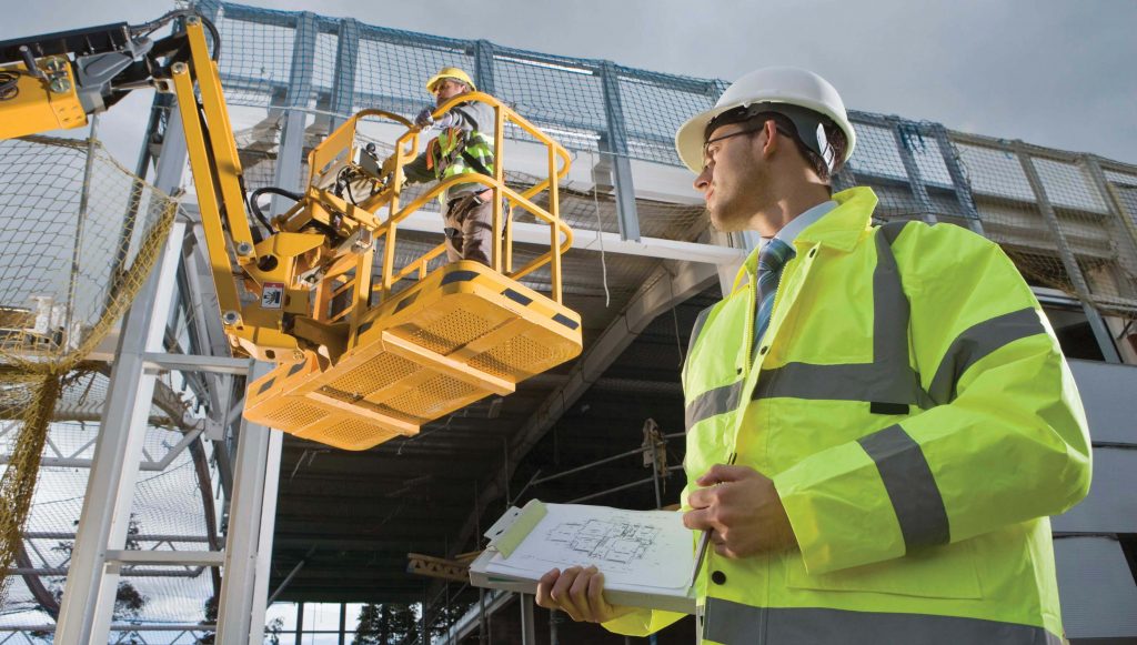 A man in a safety jacket and hard hat standing in front of a crane
