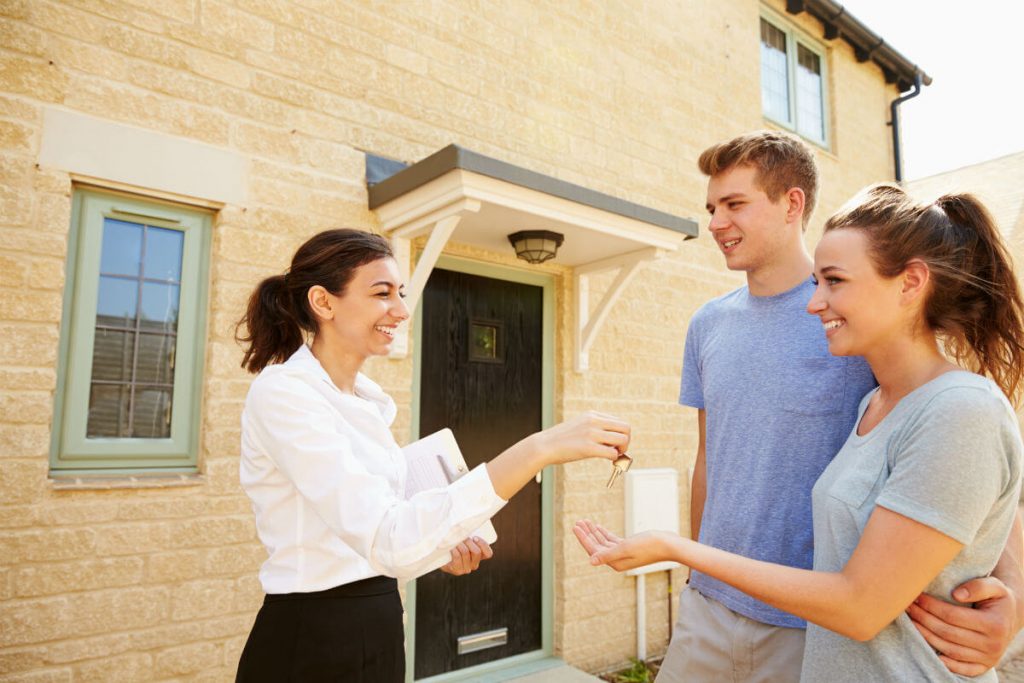 A woman handing a key to a man in front of a house
