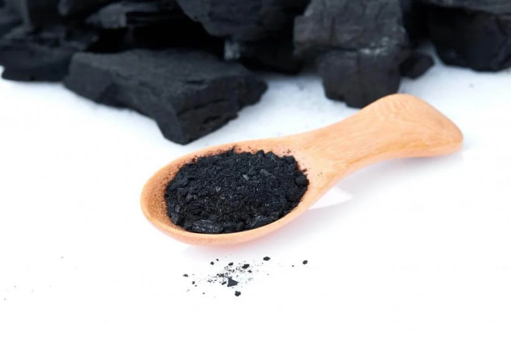 A wooden spoon filled with black Charcoal next to a pile of Charcoal

