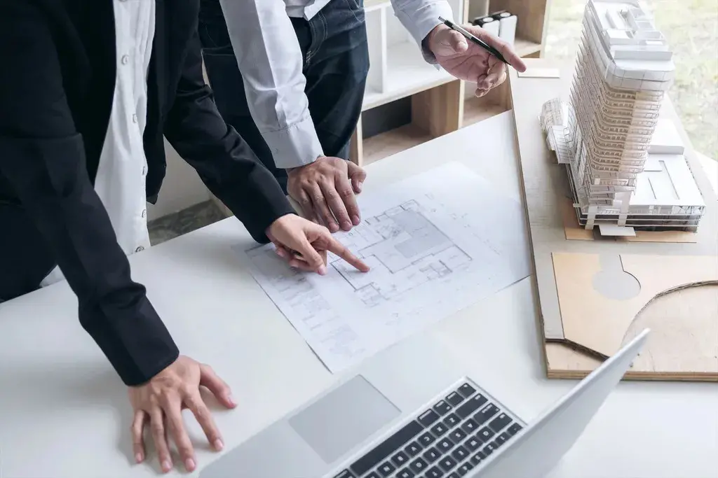 Two men in suits are pointing at a blueprint on a desk
